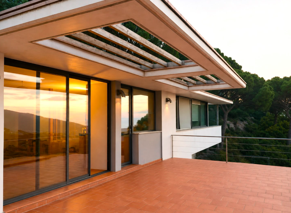 Terraza cubierta con pérgola de madera, grandes ventanales y suelo de baldosas al atardecer
