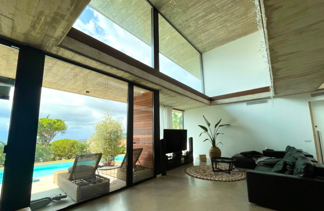 Salón a doble altura con techo de hormigón visto y ventanales hacia la piscina